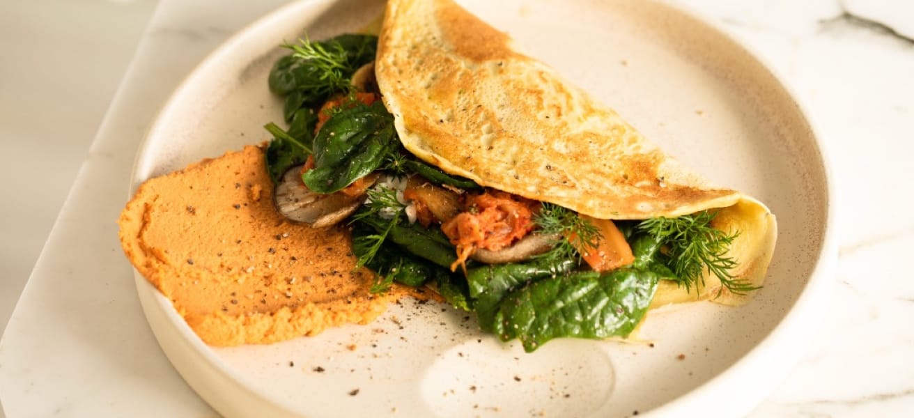 Plate with an omelette filled with sautéed vegetables and greens, garnished with herbs and served with a side of red pepper and hummus.